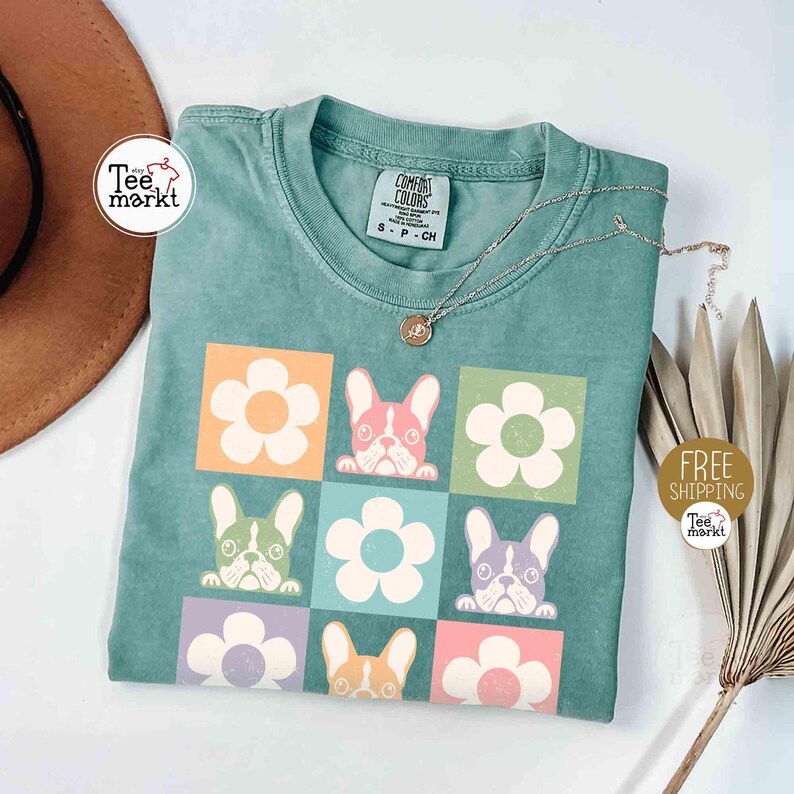 May include: A teal-colored t-shirt featuring a grid design with cartoon French bulldog faces and flower graphics in various colors. The shirt has a crew neck and short sleeves. A silver necklace with a pendant is draped across the shirt.