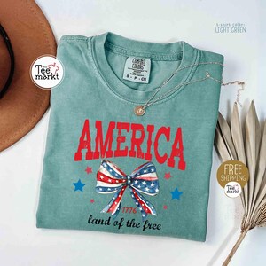May include: Light green t-shirt with the word "AMERICA" in red, a bow with a red, white, and blue pattern, and blue stars. The text "1776 land of the free" is below the bow. A silver necklace is draped across the shirt.