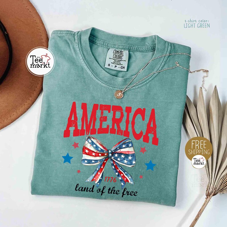 May include: Light green t-shirt with the word "AMERICA" in red, a bow with a red, white, and blue pattern, and blue stars. The text "1776 land of the free" is below the bow. A silver necklace is draped across the shirt.
