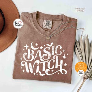 以下が含まれることがあります： 「BASIC WITCH」の白い文字が特徴の、エスプレッソ色のTシャツ。星、三日月、コウモリが描かれています。ペンダント付きのシルバーネックレスがシャツの上にあります。「COMFORT COLORS」と書かれたタグが付いています。
