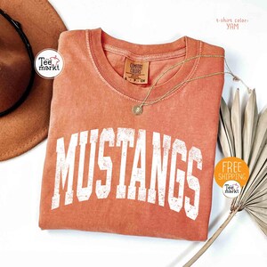 May include: A yam-colored t-shirt with the word "MUSTANGS" in large, distressed white letters. The shirt has a crew neck and a small tag. A gold necklace with a pendant is draped across the shirt.