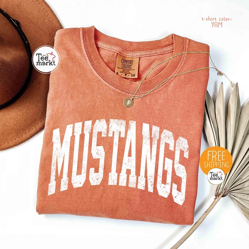 May include: A yam-colored t-shirt with the word "MUSTANGS" in large, distressed white letters. The shirt has a crew neck and a small tag. A gold necklace with a pendant is draped across the shirt.