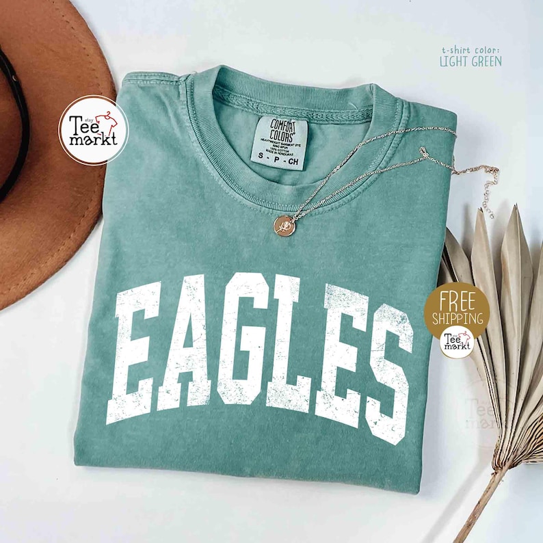 May include: Light green t-shirt with the word "EAGLES" in large, distressed white letters. The shirt has a crew neck and short sleeves. A silver necklace with a pendant rests on the shirt. The shirt is labeled "Comfort Colors".