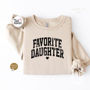 May include: A sand-coloured crewneck sweatshirt featuring the black text FAVORITE DAUGHTER in a distressed, arched varsity-style font. A small black heart icon is centred below the text. The garment has long sleeves and a ribbed collar.