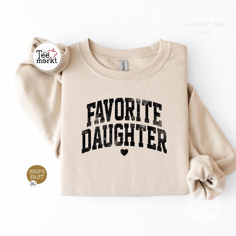 May include: A sand-coloured crewneck sweatshirt featuring the black text FAVORITE DAUGHTER in a distressed, arched varsity-style font. A small black heart icon is centred below the text. The garment has long sleeves and a ribbed collar.