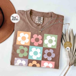 May include: Brown t-shirt with a floral design. The shirt features nine colorful flower graphics in squares. A silver necklace with a pendant rests on the shirt. The shirt has a tag that says "Comfort Colors".