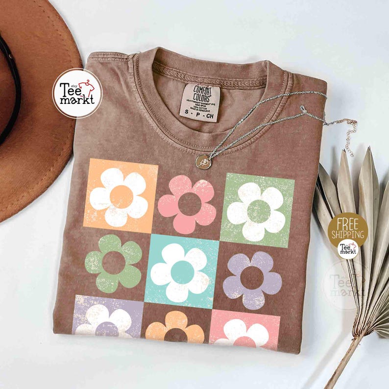 May include: Brown t-shirt with a floral design. The shirt features nine colorful flower graphics in squares. A silver necklace with a pendant rests on the shirt. The shirt has a tag that says "Comfort Colors".