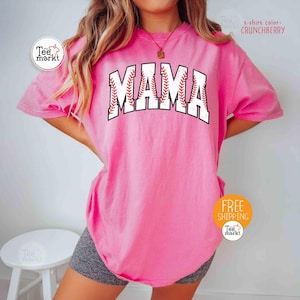 May include: A pink t-shirt featuring the word "MAMA" in a baseball design. The letters are white with red stitching, mimicking a baseball. The shirt has a solid color and a loose fit. The t-shirt colour is "Crunchberry".