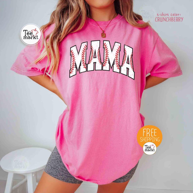 May include: A pink t-shirt featuring the word "MAMA" in a baseball design. The letters are white with red stitching, mimicking a baseball. The shirt has a solid color and a loose fit. The t-shirt colour is "Crunchberry".