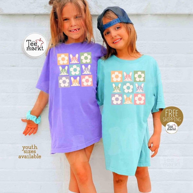 May include: Two children wearing matching t-shirts. One shirt is light purple, the other is light blue. Each shirt has a square design with flowers and bunny graphics. The text "youth sizes available" is visible.