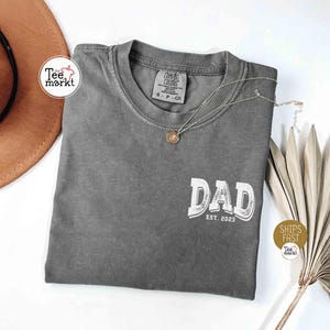 May include: A gray t-shirt with the word "DAD" in white, with "EST. 2023" below it. The shirt has a crew neck and short sleeves. A silver necklace with a pendant is on the shirt. The shirt is folded on a white surface.