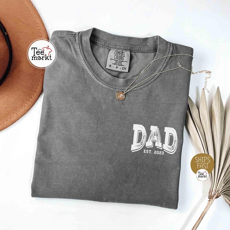 May include: A gray t-shirt with the word "DAD" in white, with "EST. 2023" below it. The shirt has a crew neck and short sleeves. A silver necklace with a pendant is on the shirt. The shirt is folded on a white surface.