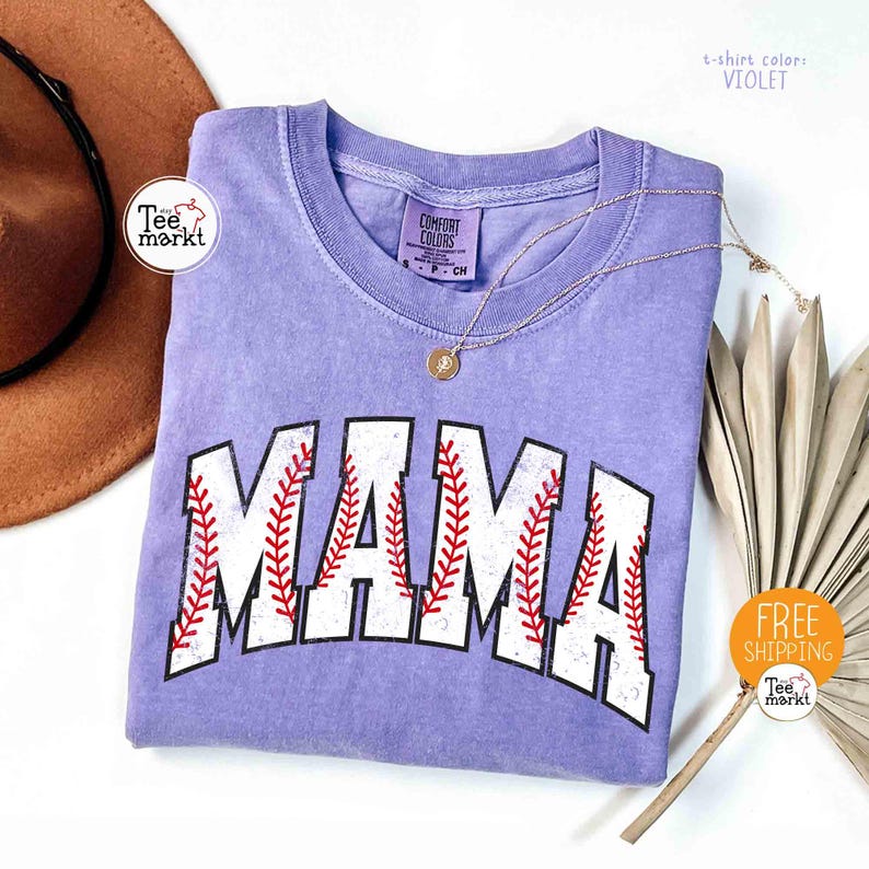 May include: A violet Comfort Colors t-shirt with the word "MAMA" in white, baseball-style lettering with red stitching. A gold necklace and a brown hat are also visible. The shirt is part of the Tee Markt collection.