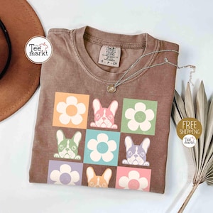 May include: Brown t-shirt featuring a colorful graphic of French bulldogs and flowers. The design includes a grid of squares with different colored backgrounds. A silver necklace with a small pendant is draped across the shirt.
