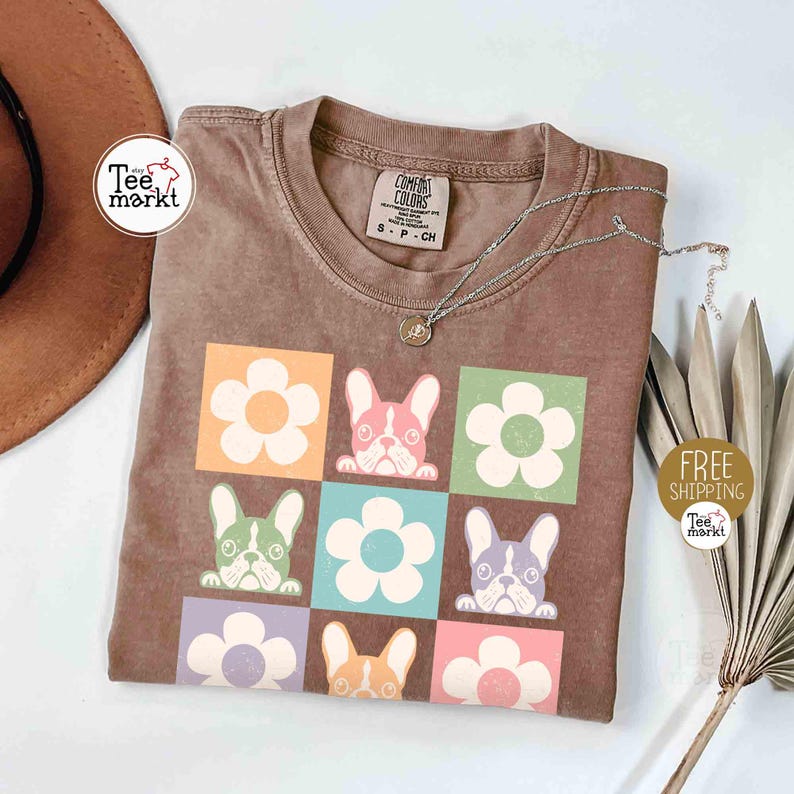 May include: Brown t-shirt featuring a colorful graphic of French bulldogs and flowers. The design includes a grid of squares with different colored backgrounds. A silver necklace with a small pendant is draped across the shirt.