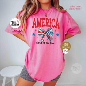 May include: A pink t-shirt with the word "AMERICA" in red block letters. Below the word is a bow in red, white, and blue, with the text "1776 land of the free". The shirt's color is "CRUNCHBERRY".