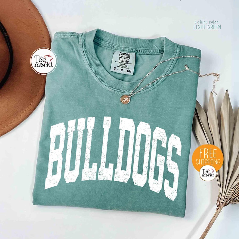 May include: Light green t-shirt with the word "BULLDOGS" in large, distressed white letters. The shirt has a crew neck and a small tag. A silver necklace with a pendant rests on the shirt.