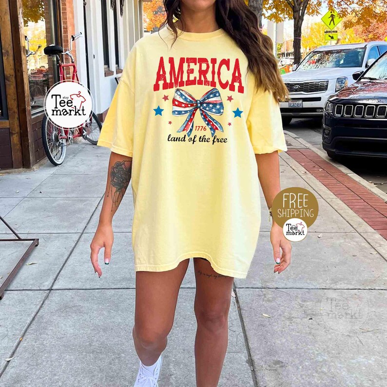 May include: A light yellow t-shirt with the word "AMERICA" in red, a bow in red, white, and blue, and the text "1776 land of the free." The shirt is a casual style.