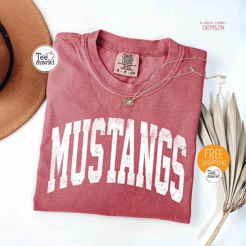 May include: Crimson-colored t-shirt with the word "MUSTANGS" in white block letters. The shirt has a crew neck and short sleeves. A gold-colored necklace with a pendant is on the shirt. The shirt is from Comfort Colors.