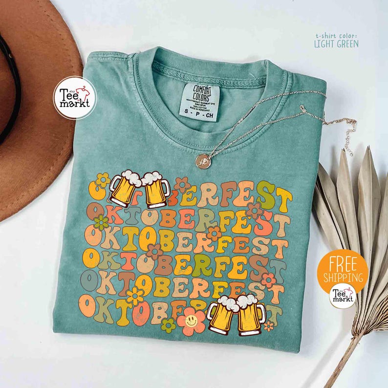 K&ouml;nnte beinhalten: Hellgr&uuml;nes T-Shirt mit dem Text "OKTOBERFEST" in einem Retro-Schriftzug, mit Bierkr&uuml;gen und Blumen. Das Shirt hat einen Rundhalsausschnitt und kurze &Auml;rmel. Eine silberne Halskette mit Anh&auml;nger liegt auf dem Shirt.