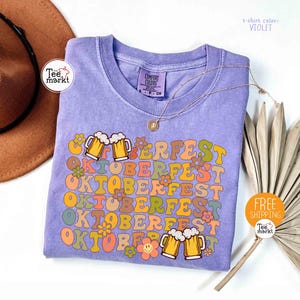 K&ouml;nnte beinhalten: Violettes T-Shirt mit dem Wort "OKTOBERFEST" in Retro-Schrift, verziert mit Bierkr&uuml;gen und Blumen. Das T-Shirt hat einen Rundhalsausschnitt und kurze &Auml;rmel. Eine goldene Halskette liegt &uuml;ber dem T-Shirt. Das T-Shirt ist von Tee Markt.