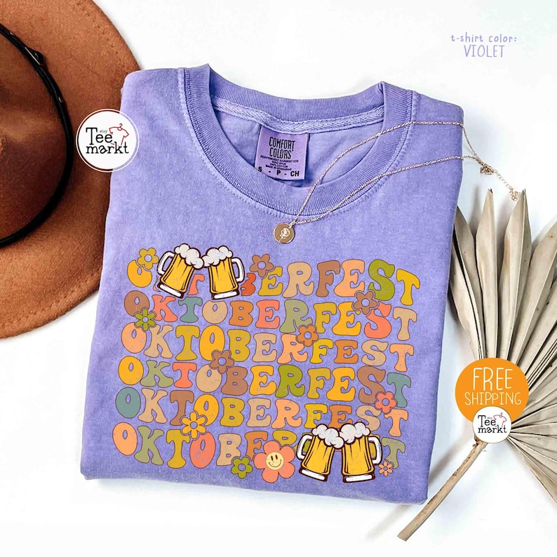 K&ouml;nnte beinhalten: Violettes T-Shirt mit dem Wort "OKTOBERFEST" in Retro-Schrift, verziert mit Bierkr&uuml;gen und Blumen. Das T-Shirt hat einen Rundhalsausschnitt und kurze &Auml;rmel. Eine goldene Halskette liegt &uuml;ber dem T-Shirt. Das T-Shirt ist von Tee Markt.