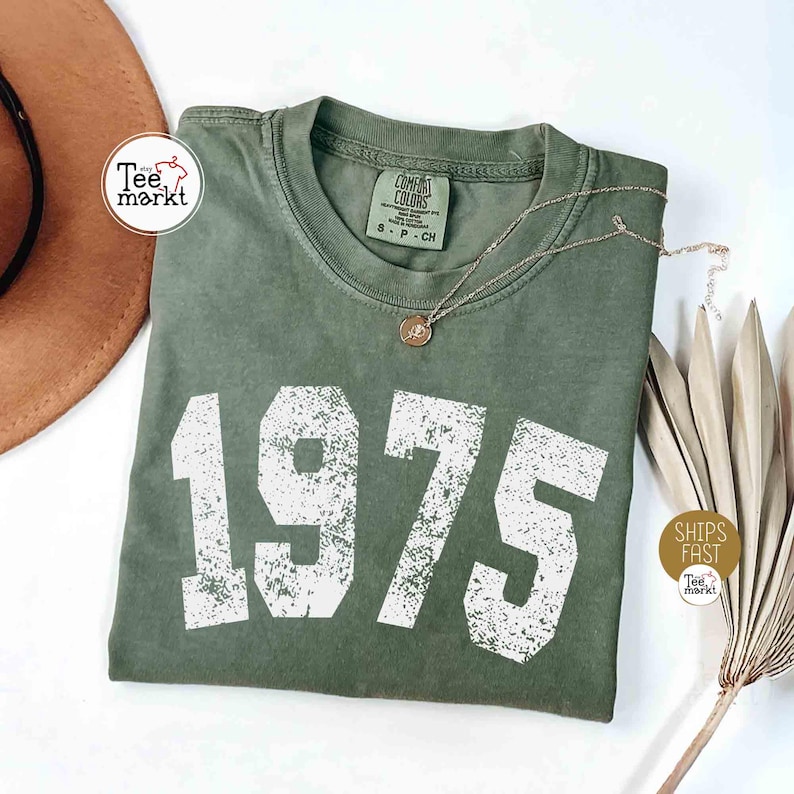 May include: A green t-shirt with the year 1975 printed in white distressed font. The shirt is folded and laid flat on a white surface. The shirt has a tag that says 'Comfort Colors' and 'S - P - CH'.