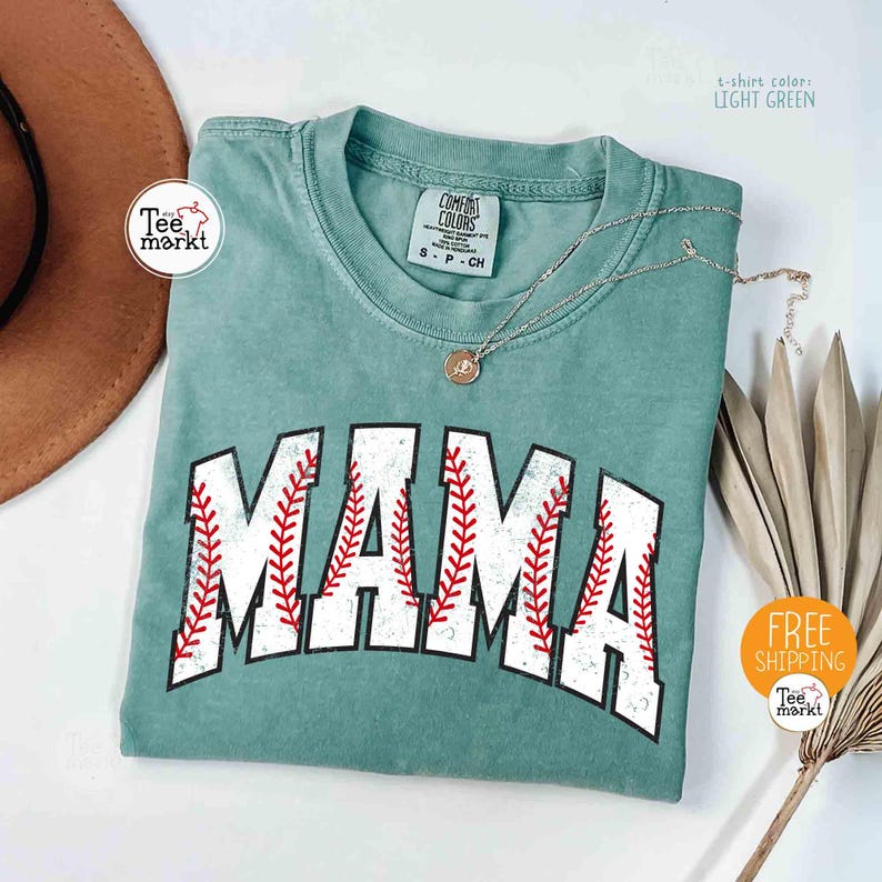 May include: Light green t-shirt with the word "MAMA" in a baseball-style font, featuring red stitching details. A silver necklace with a pendant rests on the shirt. The shirt has a tag that says "COMFORT COLORS".