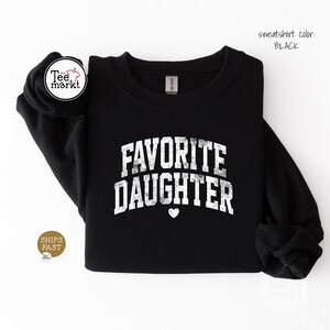 May include: A black long-sleeve crewneck sweatshirt featuring the white distressed text FAVORITE DAUGHTER in an arched collegiate font with a small white heart icon centred below the lettering. The garment has a standard ribbed neckline.
