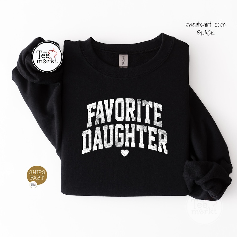 May include: A black long-sleeve crewneck sweatshirt featuring the white distressed text FAVORITE DAUGHTER in an arched collegiate font with a small white heart icon centred below the lettering. The garment has a standard ribbed neckline.