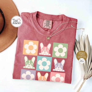 May include: A maroon t-shirt featuring a grid pattern of flowers and French bulldog faces in various colors. The shirt has a crew neck and short sleeves. A silver necklace with a pendant is on the shirt. The text "Tee Markt" is visible.