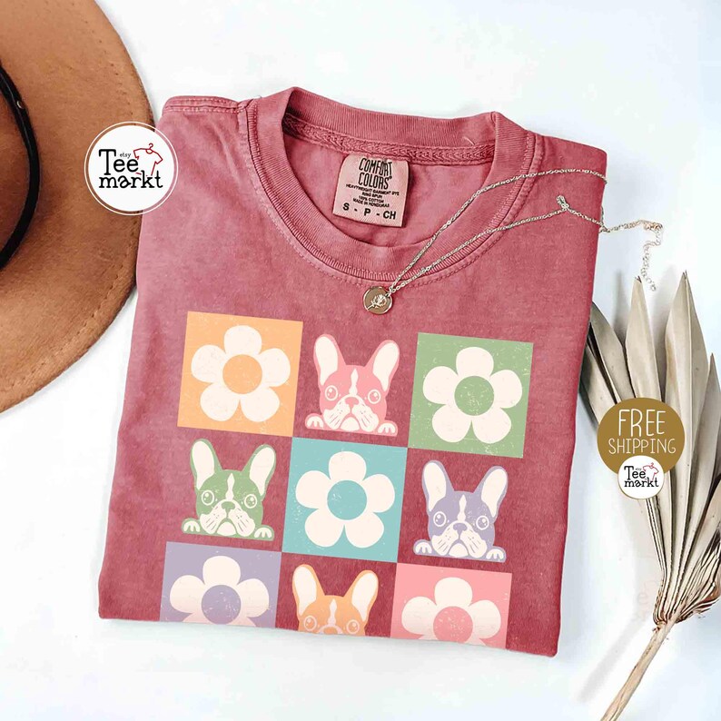 May include: A maroon t-shirt featuring a grid pattern of flowers and French bulldog faces in various colors. The shirt has a crew neck and short sleeves. A silver necklace with a pendant is on the shirt. The text "Tee Markt" is visible.