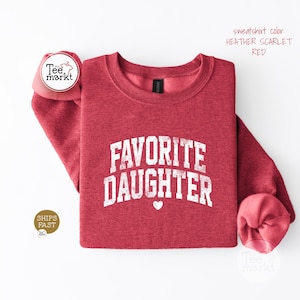 May include: A heather scarlet red crewneck sweatshirt featuring the white distressed text FAVORITE DAUGHTER in a collegiate font with a small white heart icon centred below the lettering. The garment has long sleeves and a ribbed neckline.