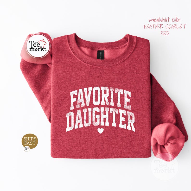 May include: A heather scarlet red crewneck sweatshirt featuring the white distressed text FAVORITE DAUGHTER in a collegiate font with a small white heart icon centred below the lettering. The garment has long sleeves and a ribbed neckline.