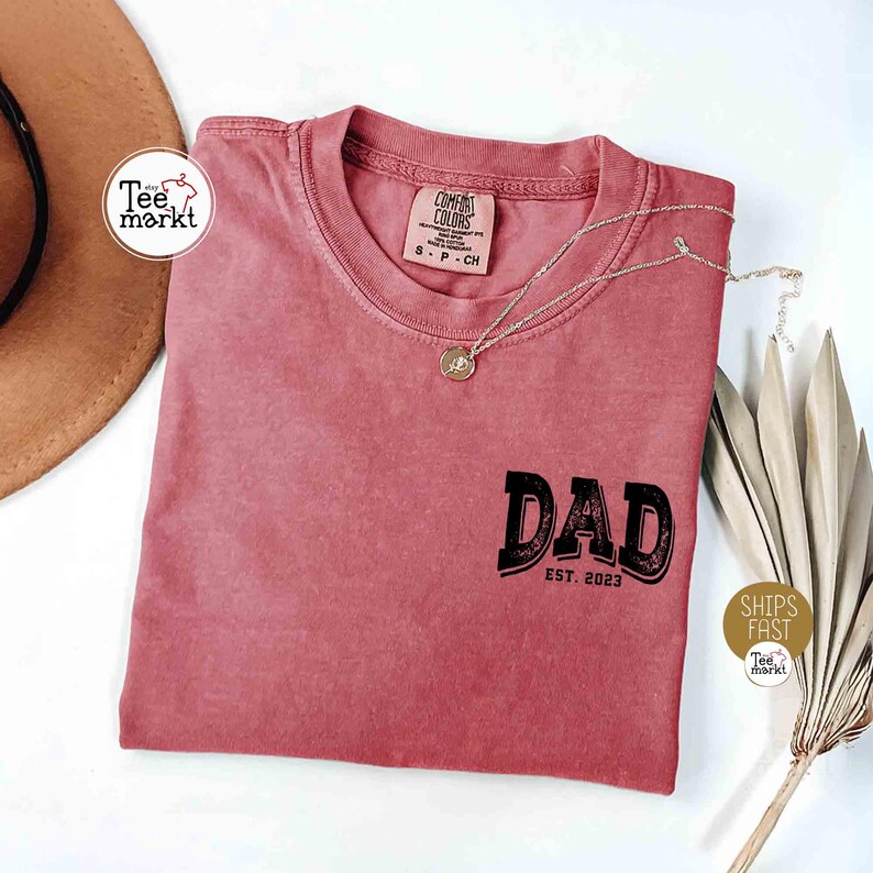 May include: A dusty rose-colored t-shirt with the word "DAD" in a black, vintage-style font, and "EST. 2023" below it. A silver necklace with a pendant rests on the shirt. The shirt has a Comfort Colors tag.