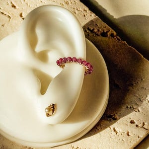 Może przedstawiać: Złota nausznica z małymi, okrągłymi, różowymi kamieniami szlachetnymi i złotą kolczykiem sztyftem z małą perłą. Biżuteria jest prezentowana na białym kształcie ucha i teksturowanej, beżowej powierzchni.