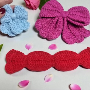 Puede incluir: Un lazo de ganchillo azul claro, un lazo de ganchillo magenta grande y una pieza de ganchillo roja larga con forma de caramelo. Un c&iacute;rculo amarillo contiene el texto Crochet Pattern PDF+VIDEO. Los objetos est&aacute;n dispuestos con una hoja verde y p&eacute;talos.
