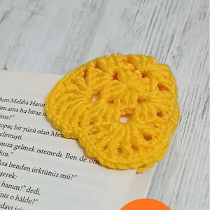 May include: A handmade, yellow crocheted bookmark in a floral design, placed on a book page. The bookmark is made of yarn and has a textured appearance. The image also includes a round orange sticker with the text "Pattern PDF + VIDEO".