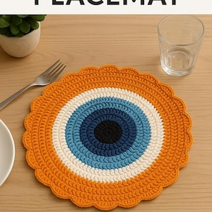 May include: A handmade, round crochet placemat with an eye design. The placemat features a scalloped edge and a central eye pattern in black, white, blue, and orange. The text "DIY CROCHET PLACEMAT" is at the top of the image.