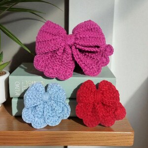 Puede incluir: Tres lazos de ganchillo hechos a mano en colores brillantes. Un lazo grande de color fucsia se sit&uacute;a sobre un lazo azul claro m&aacute;s peque&ntilde;o y otro rojo m&aacute;s peque&ntilde;o. Cada lazo presenta un dise&ntilde;o texturizado con bordes festoneados y un nudo central.