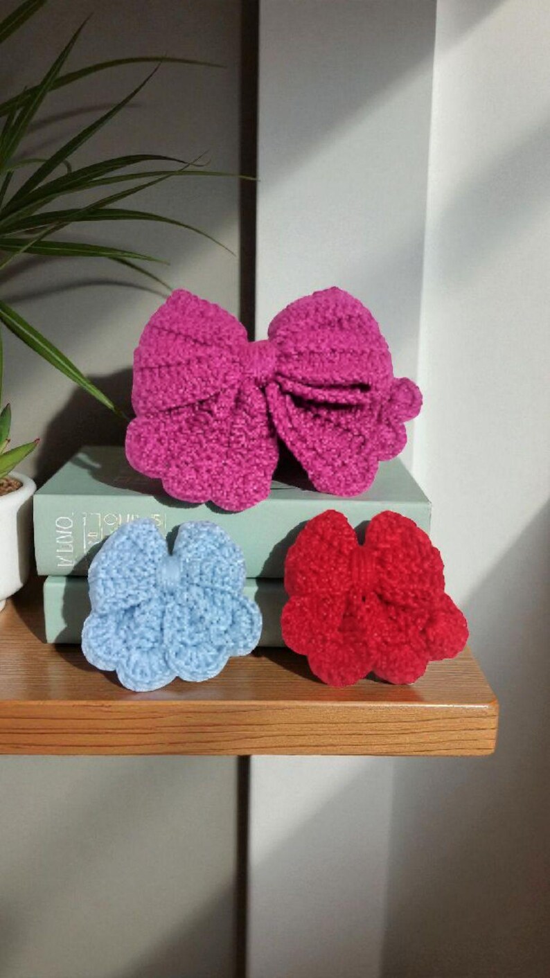 Puede incluir: Tres lazos de ganchillo hechos a mano en colores brillantes. Un lazo grande de color fucsia se sit&uacute;a sobre un lazo azul claro m&aacute;s peque&ntilde;o y otro rojo m&aacute;s peque&ntilde;o. Cada lazo presenta un dise&ntilde;o texturizado con bordes festoneados y un nudo central.