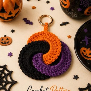 May include: A circular crochet keychain featuring three interlocking rings in orange, black, and purple yarn. The piece is attached to a gold-toned metal keyring. The text "Crochet Pattern" is printed in a cursive font at the bottom of the image.