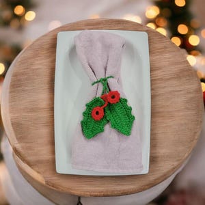 May include: A folded lavender napkin with a green crocheted holly leaf and red berry design. The napkin is placed on a white rectangular plate, which sits on a wooden surface. The design is festive.