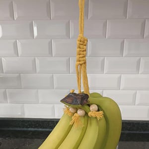 May include: A bunch of green bananas hanging from a yellow macrame banana holder. The holder has a wooden accent and decorative beads. The bananas are suspended from a metal hook.