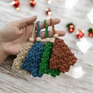 Puede incluir: Cuatro adornos de árbol de Navidad hechos a mano en beige, azul, verde y marrón. Cada árbol está hecho de hilo texturizado con un patrón de estrella y una cuenta de madera en la parte superior. Los adornos se sostienen en una mano.