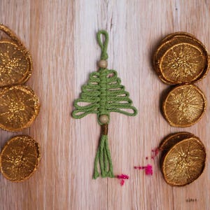 Pode incluir: Um enfeite de árvore de Natal de macramé verde feito à mão, com uma conta de madeira e borla. O enfeite é exibido com várias fatias de laranja secas pintadas de ouro em uma superfície de madeira clara. Decoração festiva.