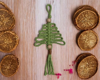May include: A handmade macrame Christmas tree ornament in green, with a wooden bead and tassel. The ornament is displayed with several gold-painted dried orange slices on a light wood surface. A festive holiday decoration.