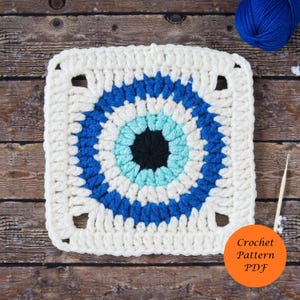May include: A square, white crochet piece with a blue and black circular design in the center. The design resembles an eye. The words "Crochet Pattern PDF" are in an orange circle in the lower right corner. A ball of blue yarn and a crochet hook are also visible.