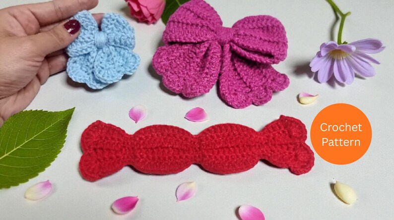 Puede incluir: Un lazo de ganchillo azul claro, un lazo de ganchillo magenta grande y una pieza de ganchillo roja larga con forma de caramelo. Un c&iacute;rculo naranja con el texto Crochet Pattern es visible. Los objetos est&aacute;n dispuestos con p&eacute;talos y hojas.