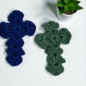 May include: Two handmade crochet crosses, one in deep blue and the other in forest green, are displayed on a white surface. The crosses have a detailed, textured design. A small potted succulent plant is in the background.